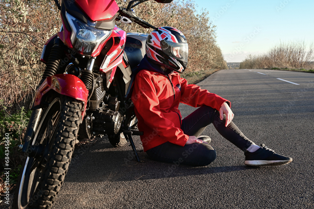 Obraz premium young attractive biker girl in red sitting next to her red motorcycle in the middle of the road. biker with a motorcycle outside the city.