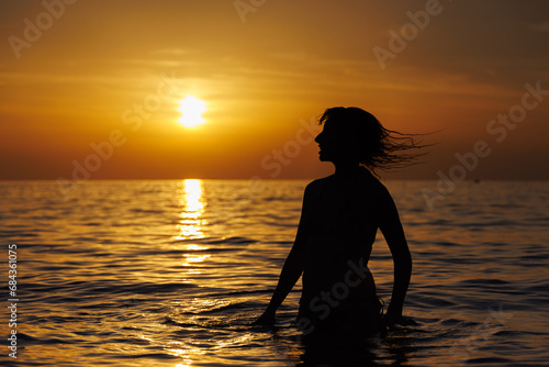 Silhouette of a girl with flying hair at sunset stands in the sea.