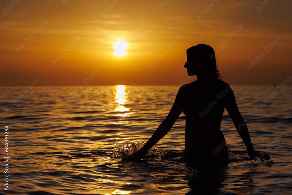 Silhouette of a girl looking at the sun and touching the sea at sunset.
