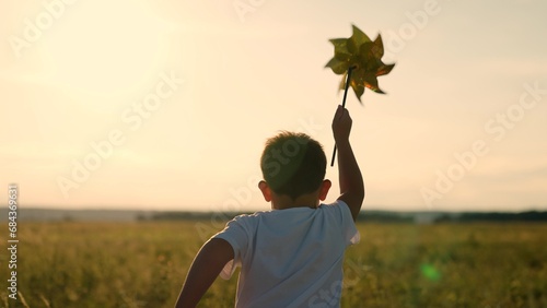 Happy child playing with toy pinwheel outdoors in summer in park against blue sky. Child boy runs with toy wind turbine in his hand on summer field, sun day. Childhood children. Family holiday, nature photo