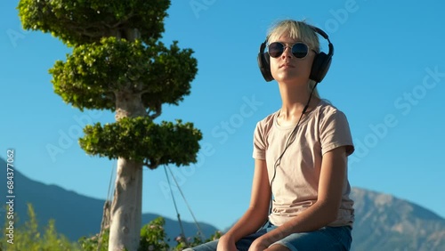 Listening to music against tropical park. A view of young girl relax on park and listening the music in headphones. photo