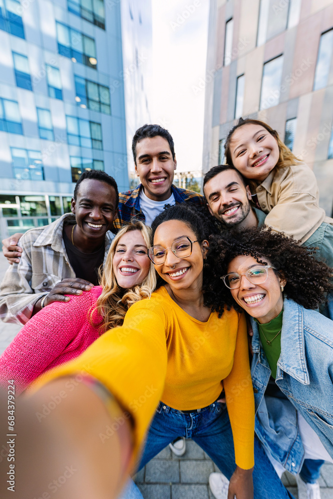 © Xavier Lorenzo - Young adult diverse friends smile at phone camera on selfie group portrait. Teenage community concept with joyful millennial students having fun together at city street.