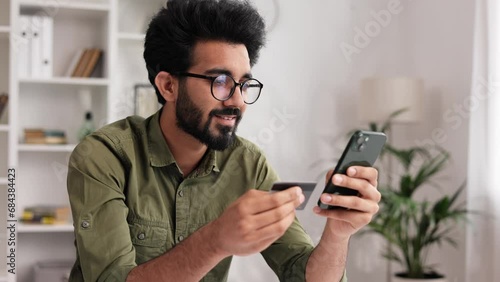 Smiling pretty young Indian brunette boy holding mobile phone and credit card sitting at the table. Happy man makes online shopping using mobile payment. E-banking app service.
