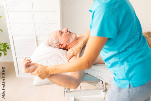 An older man, lying on the table in a physiotherapy clinic, during a manual therapy session with his physio, which assesses shoulder mobility