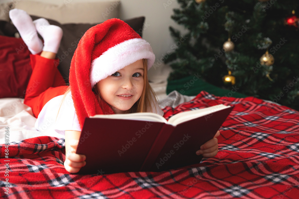Child in bed read a book. Happy little smiling girl in pajamas and Santa hat with gift on xmas lies on bed. child holding book in hand and reading it,, reads interesting fairy tale. Child in bed read a book. Happy little smiling girl in pajamas and Santa hat with gift on xmas lies on bed. child holding book in hand and reading it,, reads interesting fairy tale.