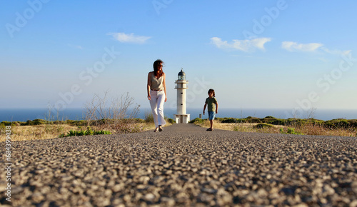 Mother and son walking towards Cap de Barberia's lighthouse, which stands beautiful on background, Formentera, Balearic Islands, Spain.