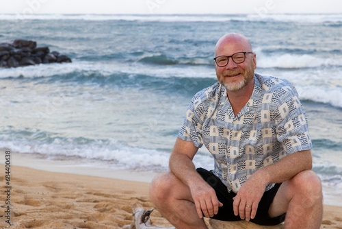 Friendly bald man with glasses and a beard wearing shorts. The middle aged guy is smiling while sitting on a log on a tropical island beach in Hawaii. He is on vacation and relaxed.  photo