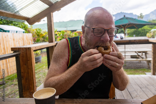 Bald man wearing glasses and a tank top tee shirt aggressively eating a hot donut outside.