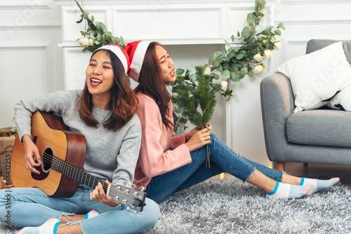 Happy Asian Woman sitting Christmas gift box under xmas tree playing acoustic guitar together. Two Women smile talk sitting decor christmas ball tree xmas holiday winter festival in living room