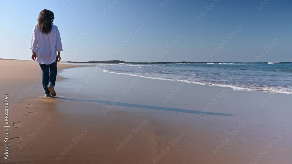 Rear view of ethnic woman in white shirt and blue jeans, enjoys her weekend outdoors, breathing fresh Atlantic air while walking and leaving footprints on the beautiful wet sandy beach. Copy ad space Rear view of ethnic woman in white shirt and blue jeans, enjoys her weekend outdoors, breathing fresh Atlantic air while walking and leaving footprints on the beautiful wet sandy beach. Copy ad space