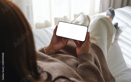Mockup image of woman holding mobile phone with blank desktop white screen while lying on bed