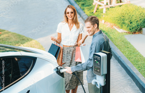 Young couple travel with EV electric car charging in green sustainable city outdoor garden in summer shows urban sustainability lifestyle by green clean rechargeable energy of electric vehicle innards photo