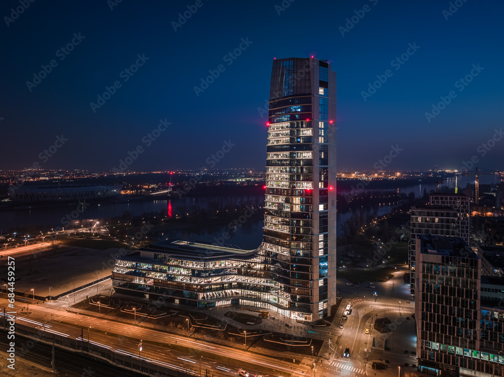 Fototapeta premium Budapest, Hungary - Aerial view of Budapest's new, illuminated skyscraper building with National Athletics Center and city skyline at background at dusk with clear blue sky