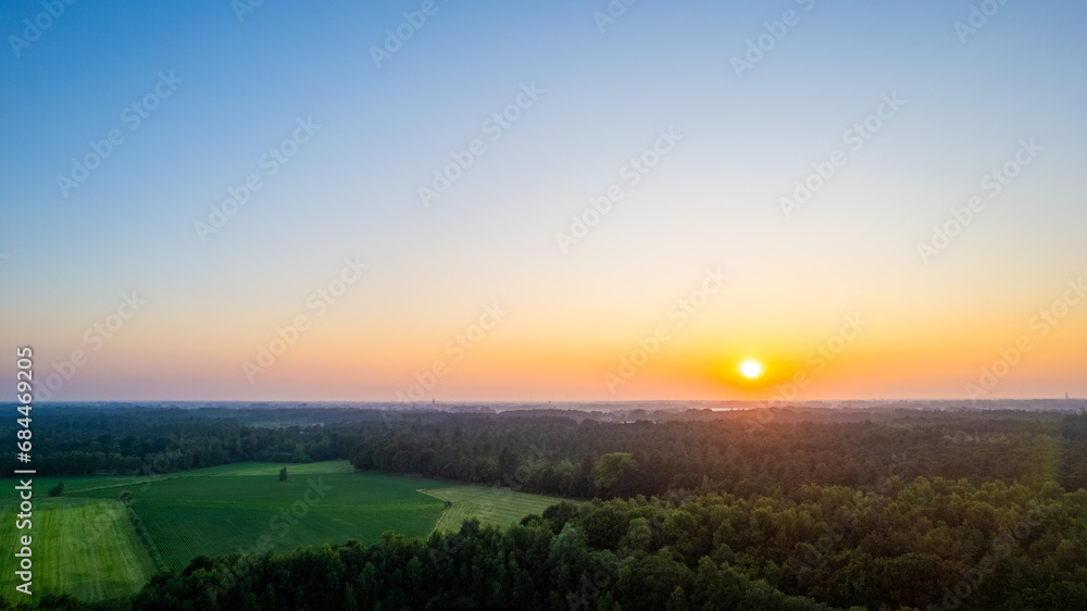 The image captures the serene beauty of sunrise over a tranquil rural landscape. The horizon is ablaze with the soft, warm hues of the early morning sun, as it rises above a dense forest and casts its