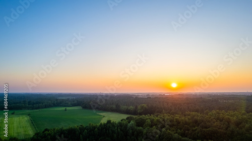 The image captures the serene beauty of sunrise over a tranquil rural landscape. The horizon is ablaze with the soft, warm hues of the early morning sun, as it rises above a dense forest and casts its