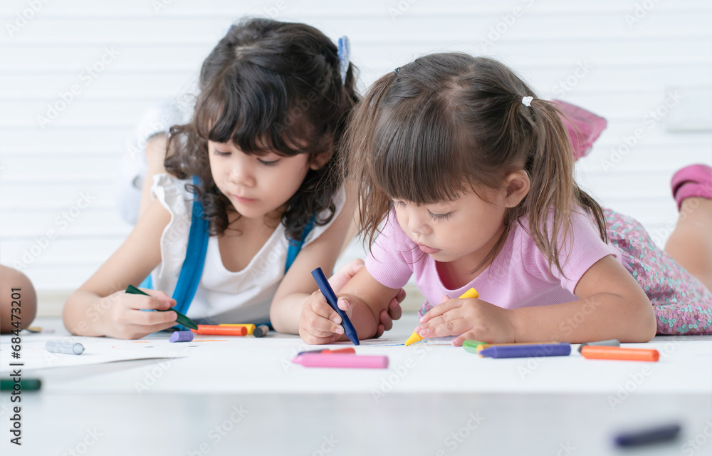 Cute little kid girl concentrate with using two hands drawing on paper while lying on floor with friends. Adorable happy children drawing with colorful crayons at school. Education, creativity concept