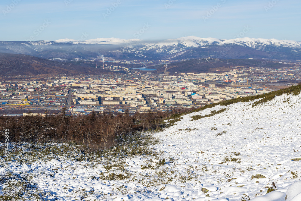 View from the mountain to the city of Magadan. In the foreground is a snow-covered hillside. In the distance there is a city and snow-capped mountains. Northern nature. Magadan, Magadan region, Russia