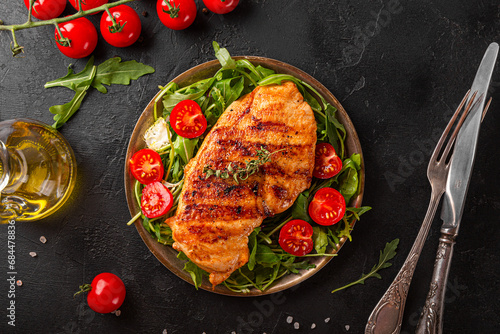 Grilled chicken breast with arugula and tomatoes on a plate, top view, black food background photo