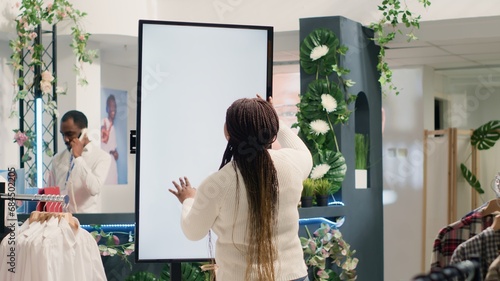 African american woman in fashion boutique using mockup augmented reality screen to look at clothes options to try on. Customer using led kiosk to visualize outfit combinations in store before buying photo