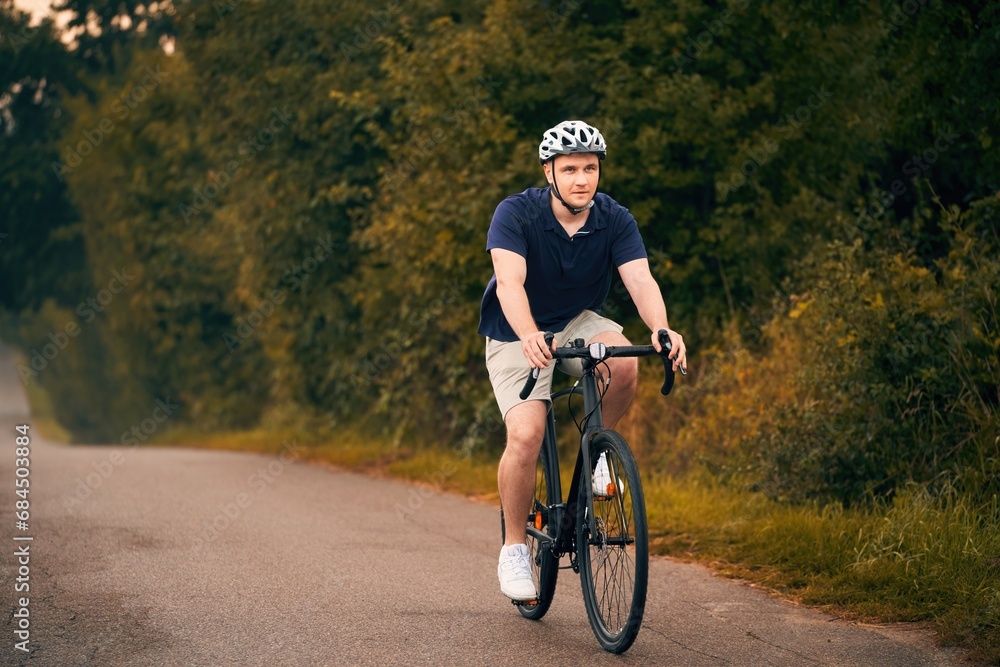 Obraz premium A young and fit cyclist on a bicycle explores the country landscape, riding on a road in the forest with a helmet and a mountain bike. Gravel biker on a path, enjoying the sport and the nature.