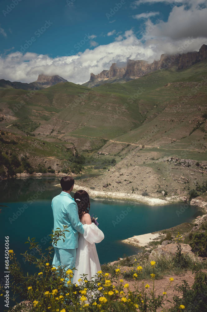 Naklejka premium Bride and groom dressed elegant in their wedding gowns holding hands and hugging posing for their photo session in a desert like landscape by the lake