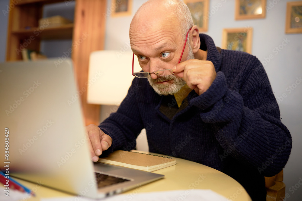 Aging and eyesight problems. Side view image of senior bald bearded grandfather in elegant stylish blue cardigan using laptop in his cabinet, reading article, adjusting glasses on his nose Aging and eyesight problems. Side view image of senior bald bearded grandfather in elegant stylish blue cardigan using laptop in his cabinet, reading article, adjusting glasses on his nose