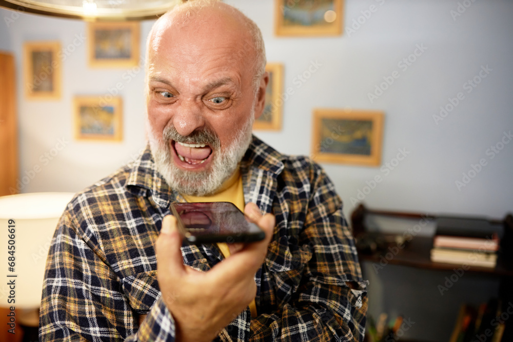 Senior man in checkered shirt yelling at smartphone. Angry furious bald bearded man recording voice message, expressing rage and displeasure, shouting with opened mouth, scolding someone - obrazy, fototapety, plakaty 