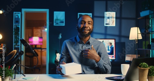 Tech content creator filming packaged product unboxing in neon lights personal home studio. BIPOC cheerful influencer showing sponsoring brand merchandise, urging subscribers to purchase