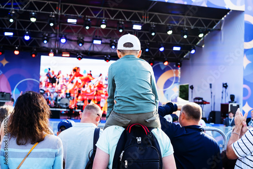 A boy sits on his dad's shoulders during a street concert. A father holds his son in a crowd of people during an open-air festival. Entertainment program of a traveling theater or a musical group