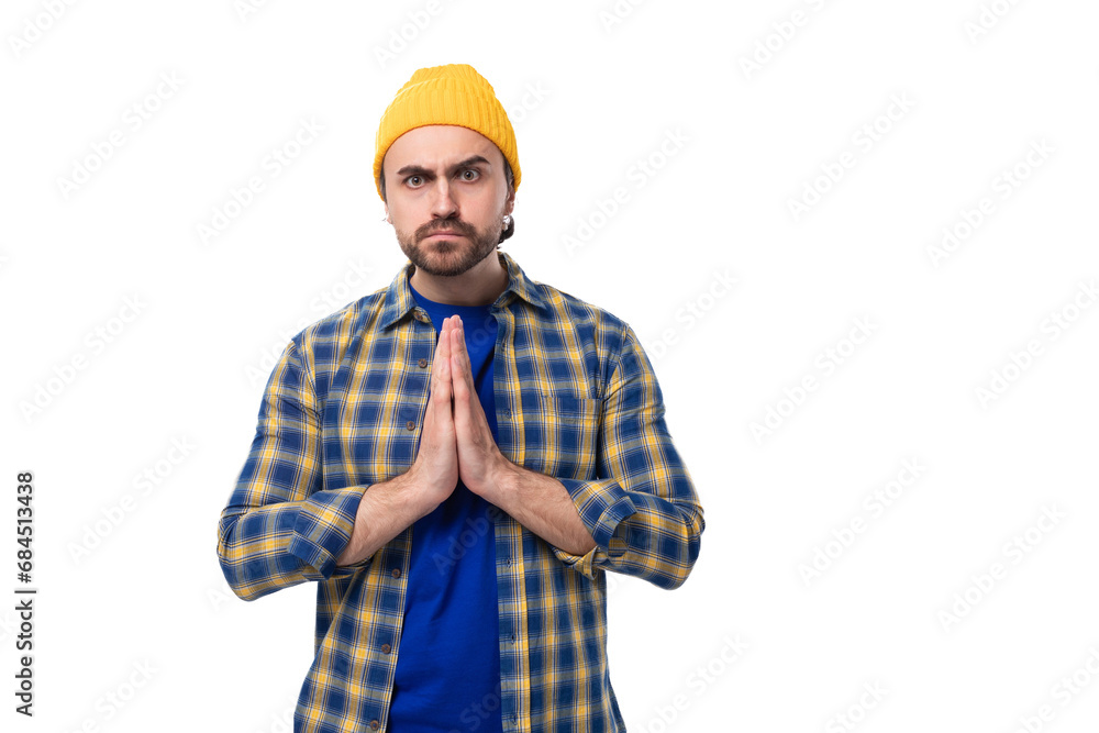 well-groomed young european brunette man with a beard and mustache dressed in a yellow hat and blue shirt put his hands together in hope