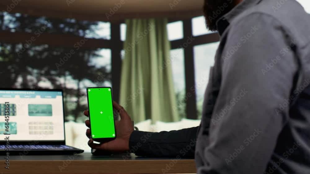 Male billionaire uses greenscreen phone display at desk in his opulent alpine retreat, rich entrepreneur at vacation home. Young shareholder works with isolated chromakey, investing capital funds. Male billionaire uses greenscreen phone display at desk in his opulent alpine retreat, rich entrepreneur at vacation home. Young shareholder works with isolated chromakey, investing capital funds.