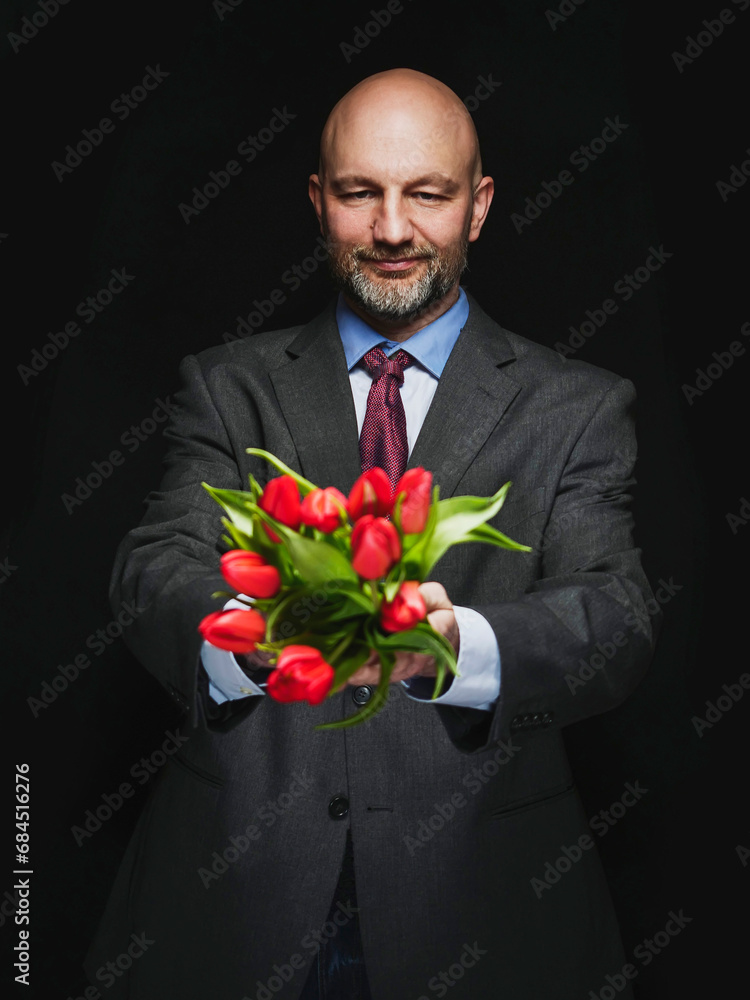 Man holding fresh red tulip bouquet in his hands. Model is bald with grey beard, wearing classic grey suit. Handsome male with special gift. Expression of feelings . Dark color background. Man holding fresh red tulip bouquet in his hands. Model is bald with grey beard, wearing classic grey suit. Handsome male with special gift. Expression of feelings . Dark color background.