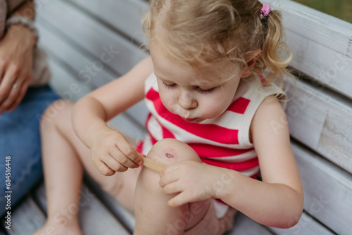 Little girl blowing on injured knee and putting on band aid photo