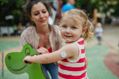 Mother watching little girl with a continuous glucose monitor sitting on dinosaur seesaw on the playground