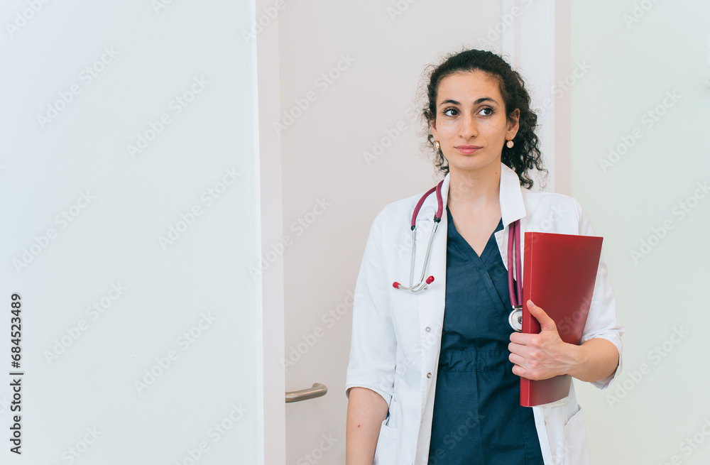 Brunette medical female employee entered office at clinic dressed in medical gown, with phonendoscope holds folders. Young doctor standing at hospital against wall with copy space for ad. Brunette medical female employee entered office at clinic dressed in medical gown, with phonendoscope holds folders. Young doctor standing at hospital against wall with copy space for ad.