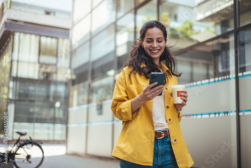 Obraz na plátně Concentrated female pedestrian in stylish outfit text messaging on mobile phone while walking along pavement near contemporary building