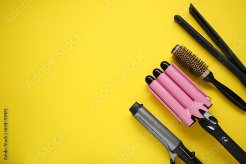 Different curling irons, hair straightener and round brush on yellow background, flat lay. Space for text photo