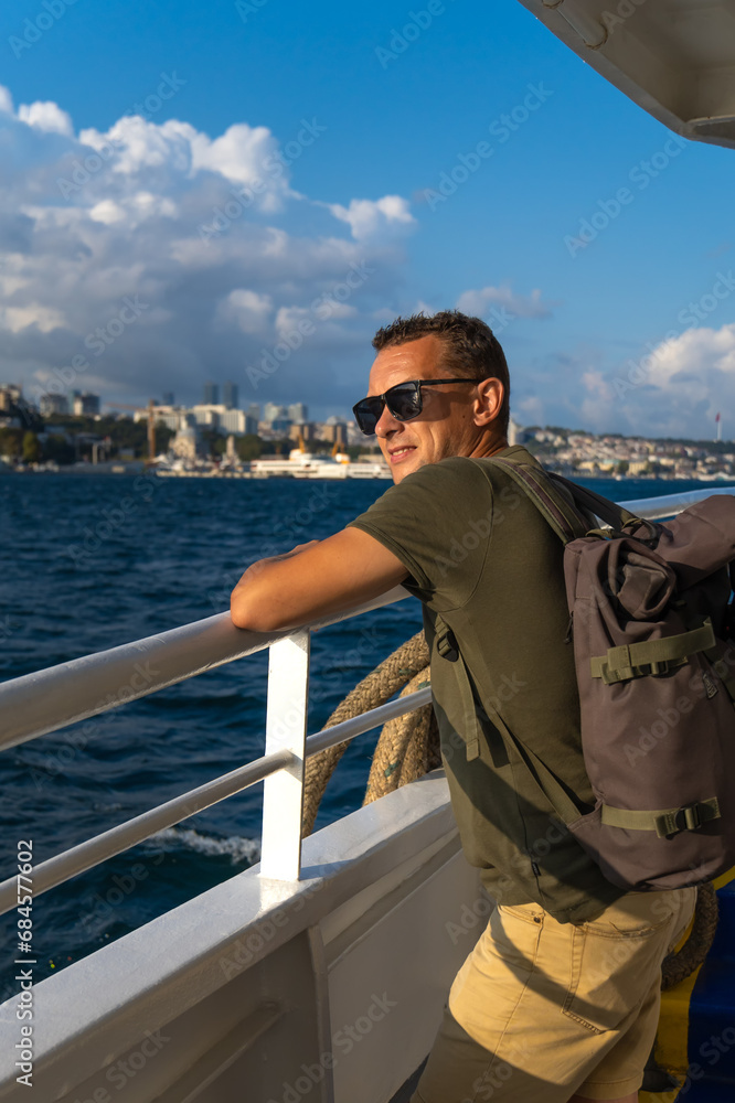 Happy handsome young male tourist in sunglasses with a backpack on his shoulders enjoys the sea and the city from a ferry in Istanbul on a sunny day. Concept of travel, tourism. Vertical photo