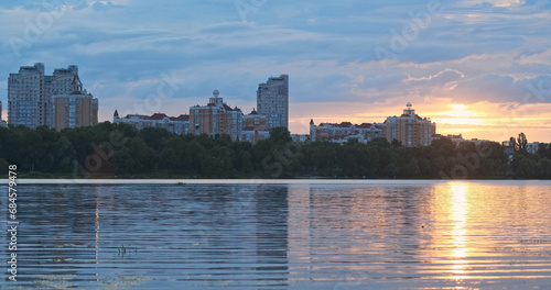 Sunset urban outskirts. Wide river, reflection sky water, sun's rays among clouds. High-rise residential buildings. Background