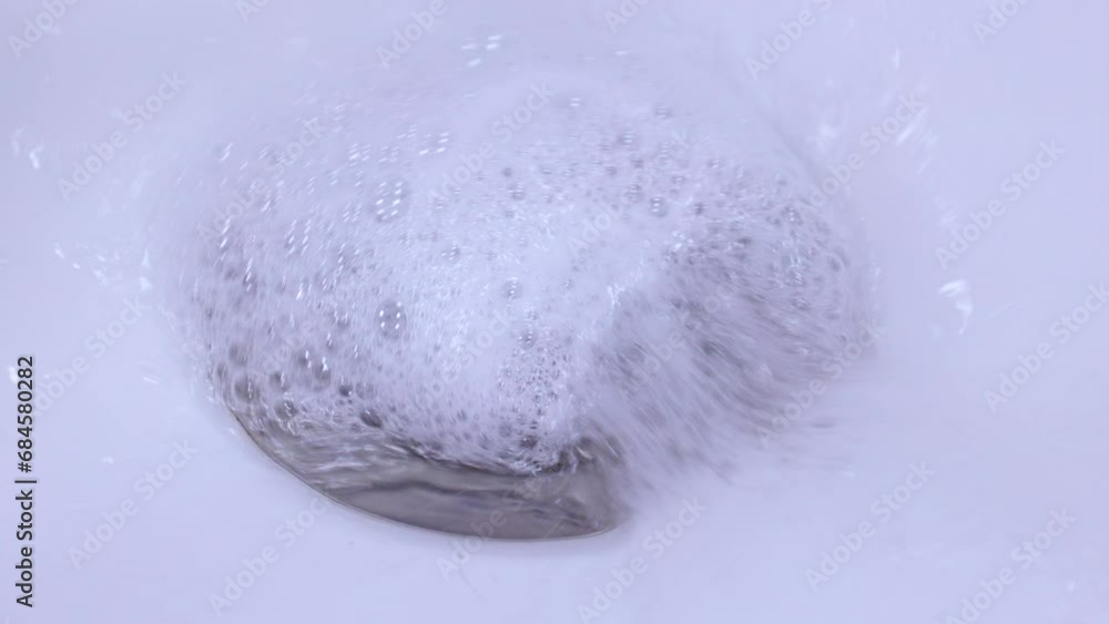 Metallic drain hole in the ceramic sink with flowing water with white soap foam in the bathroom close up Metallic drain hole in the ceramic sink with flowing water with white soap foam in the bathroom close up