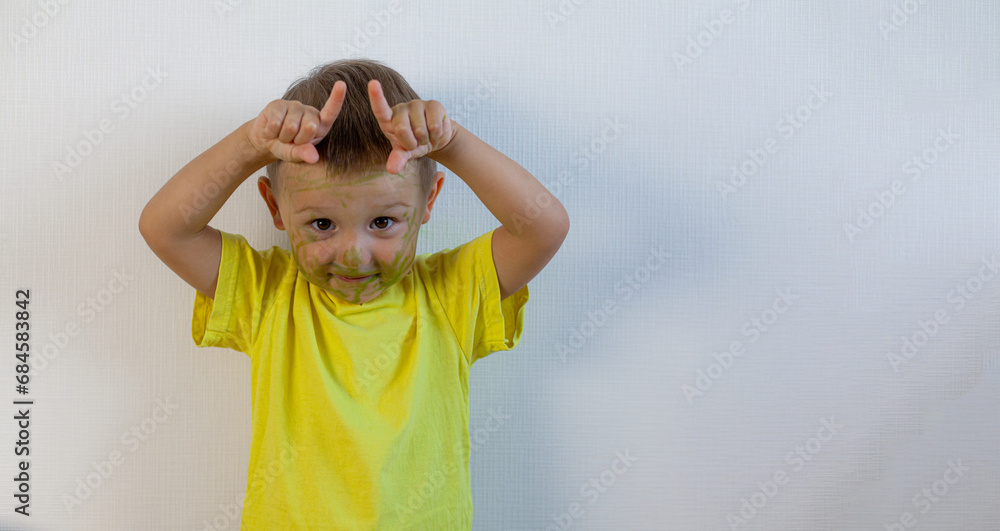 A 3-year-old boy painted his face with a marker. The boy stands at the white wall and makes horns. Banner with space for text A 3-year-old boy painted his face with a marker. The boy stands at the white wall and makes horns. Banner with space for text