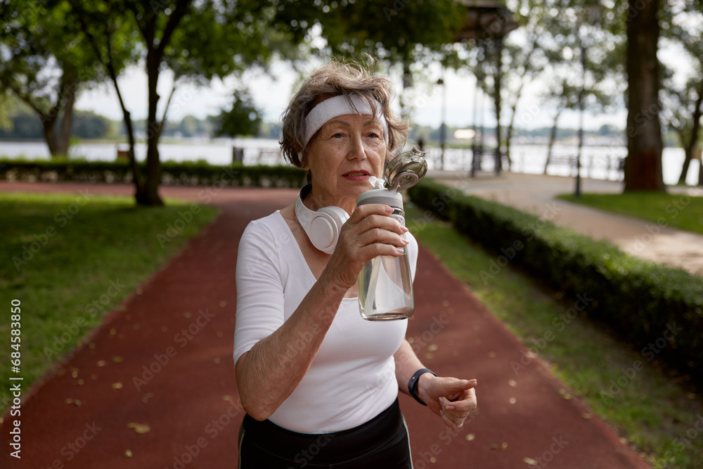 Fototapeta premium Elderly woman drinking water taking short break during running exercise
