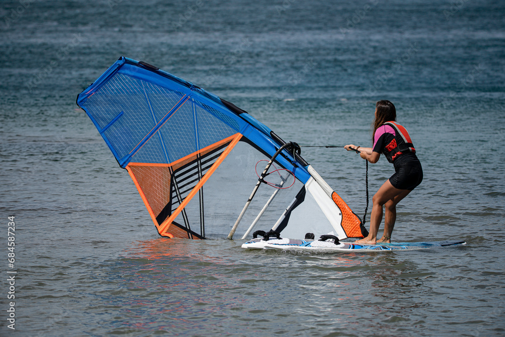 Naklejka premium young sporty girl on the windsurf board raising the sail pulling the rope