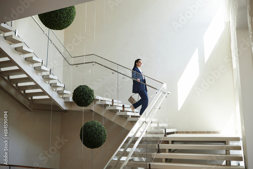 Wide angle shot of Caucasian woman in business suit carrying purse going downstairs holding on handrail in modern office building, copy space photo