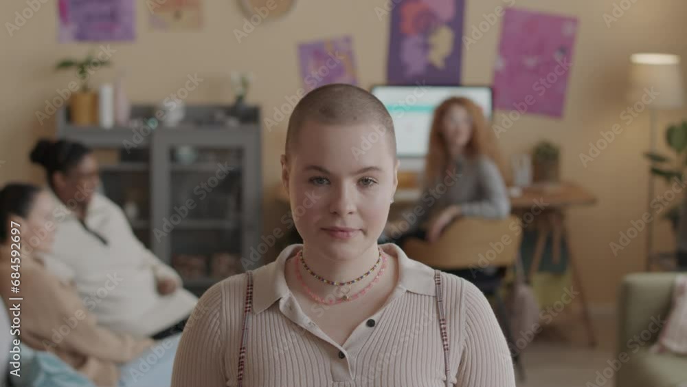 Chest up slowmo portrait of young Caucasian woman with shaved head looking confidently at camera while standing in modern studio apartment with posters of feminism symbols on walls Chest up slowmo portrait of young Caucasian woman with shaved head looking confidently at camera while standing in modern studio apartment with posters of feminism symbols on walls