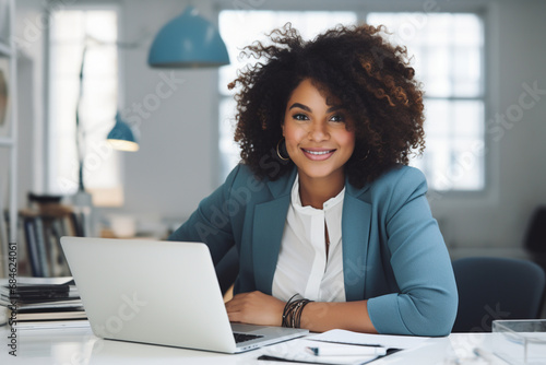 charming plump black woman of plus-size, manager, in blue business clothes sits at a desk with a laptop in a modern elegant office and smiles sweetly, the concept of diversity photo