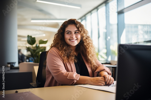 a plump woman of plus-size, a manager, in pink business clothes sits at a table in front of a monitor and smiles sweetly in a modern office, the concept of diversity