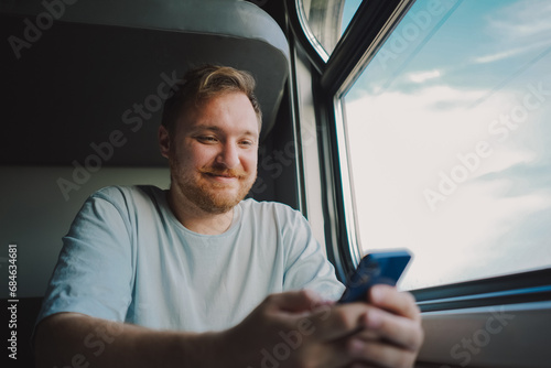 A man with a beard and mustache in a blue t-shirt is using a smartphone while traveling by Railway train, sitting in the train and looking out the window. © Анастасія Стягайло