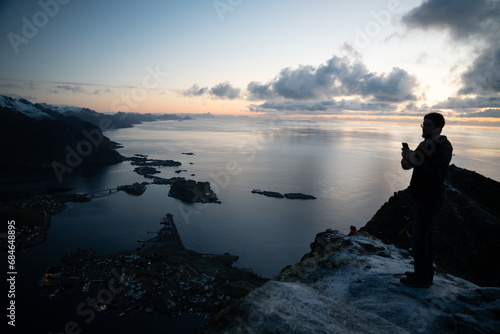 Man taking picture at top of Reinebringen mountain hike in Lofoten  Norway.  Snow covered mountains captured on image by Sony mirrorless camera.  Located far North in the Arctic Circle.  A famous hike