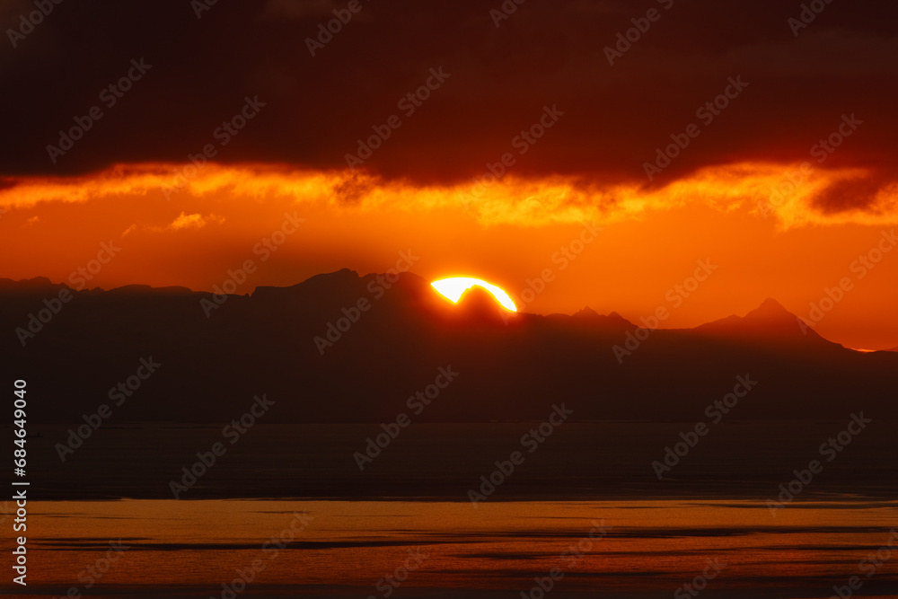 Telephoto image of large setting sun overtop a fjord mountain in Norway.  Beautiful orange sun rising up from the peaks of Norwegian mountains.  Shot from the top of Reinebringen